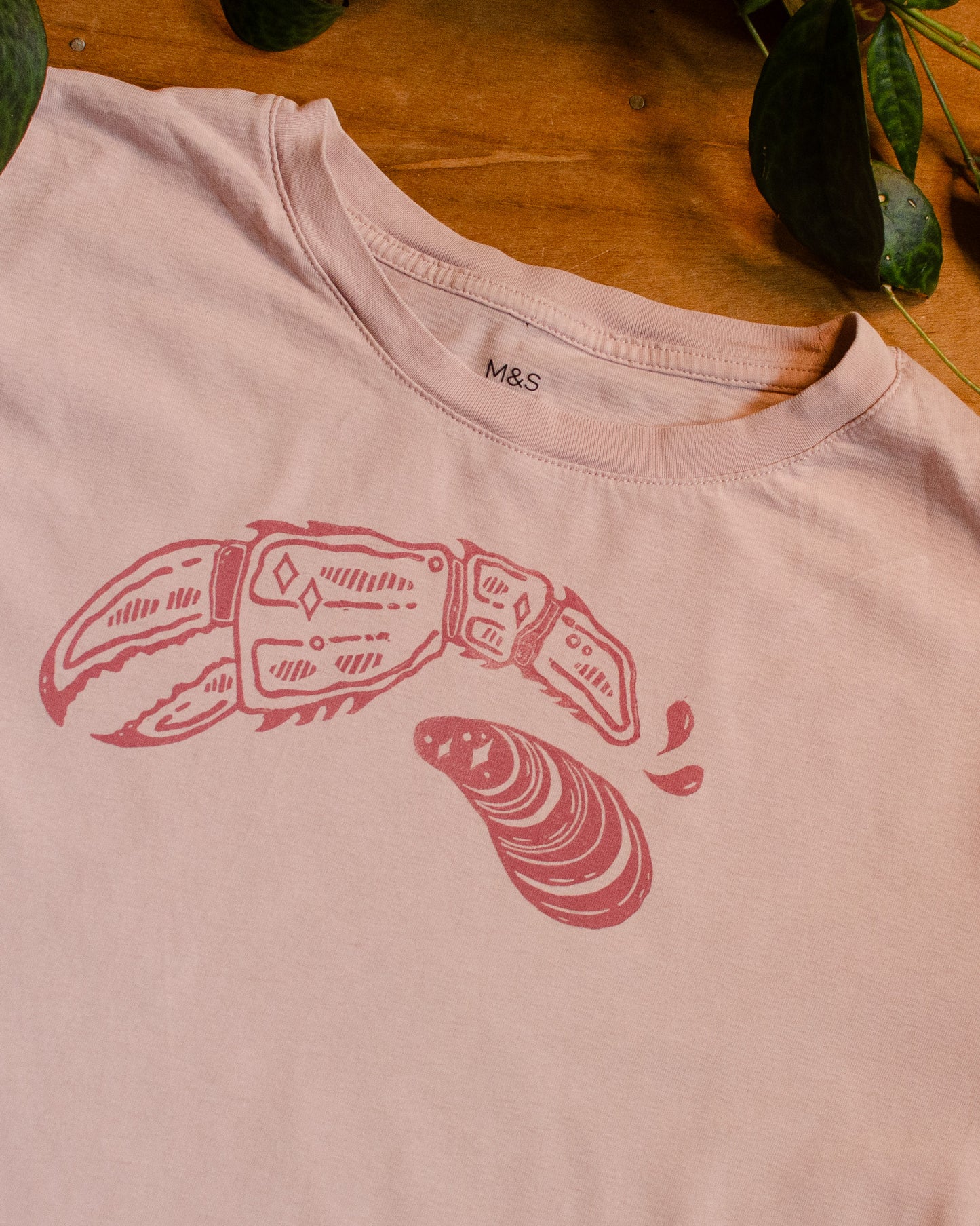 Pink t-shirt with dark pink lobster claw and mussel design on a wooden surface