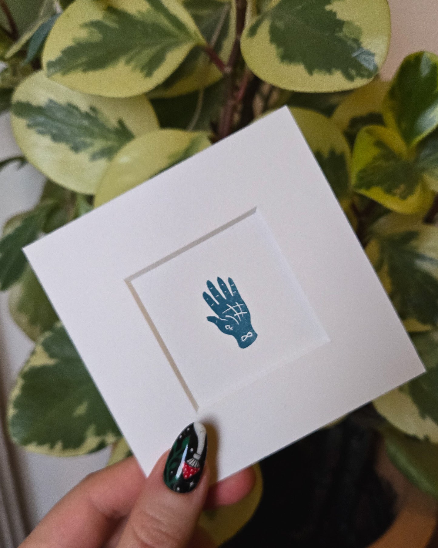 White card with a blue hand design held by a person with green nail polish against a leafy background