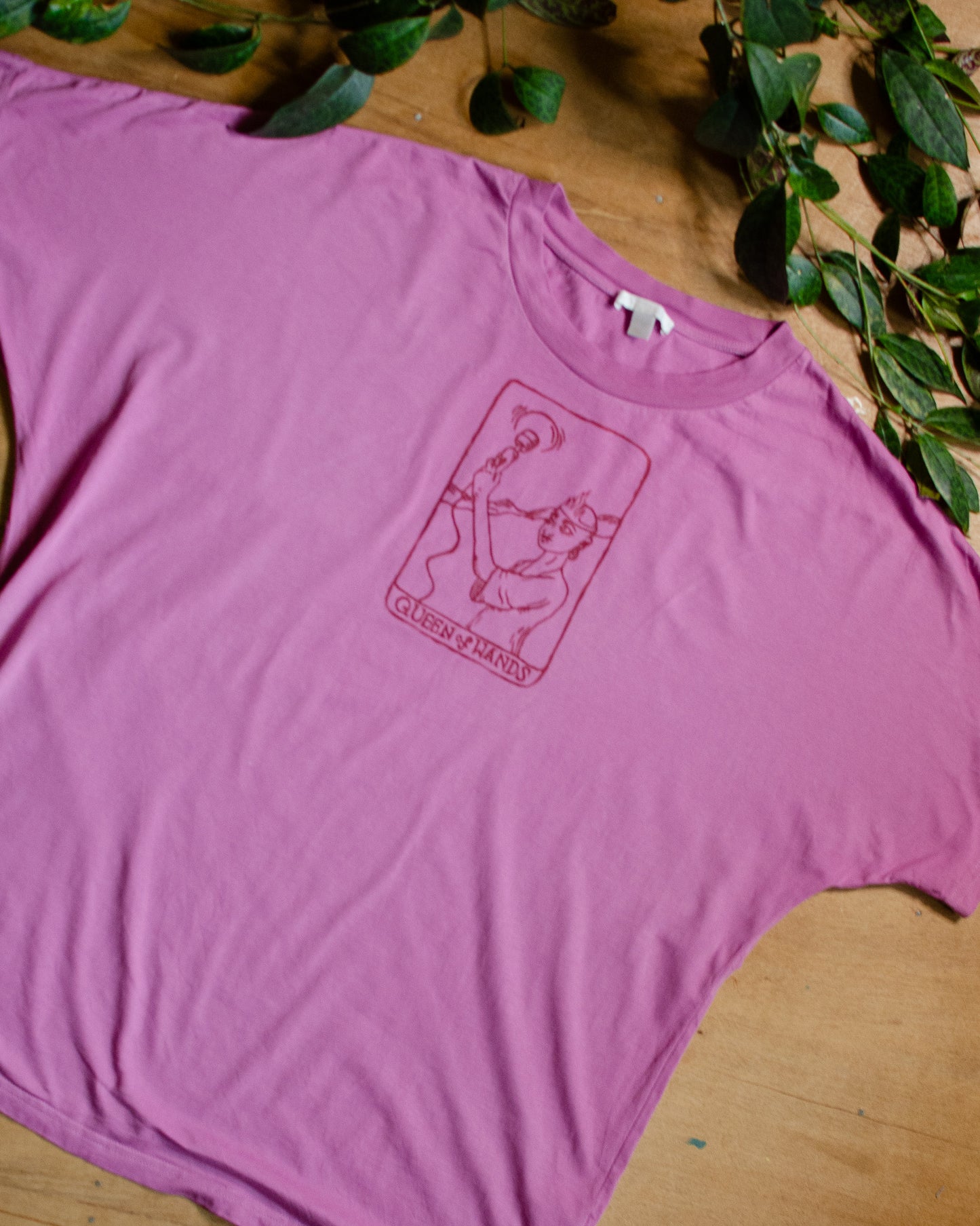 Pink t-shirt with a queen of wands tarot card design on a wooden surface with green leaves.