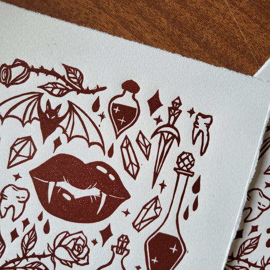 Linocut print of a vampire witch design on white paper, with bats, blood red lips, crystals, roses, and potions.