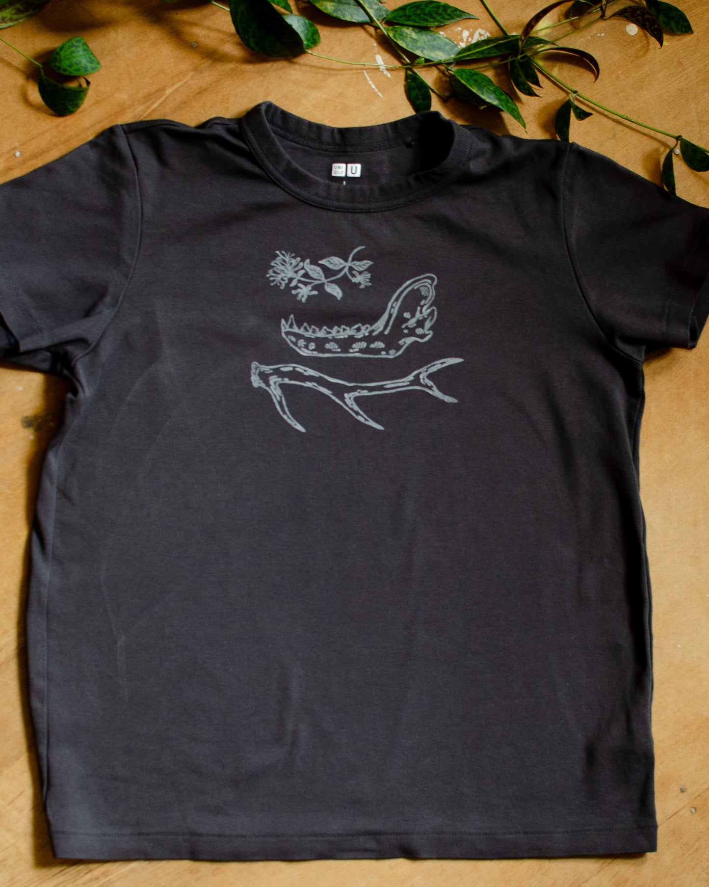Black t-shirt with hand printed white honeysuckle, jawbone, and antler design on a wooden surface with green leaves.