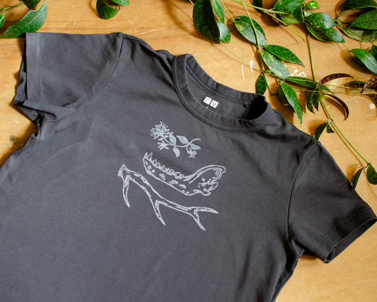 Black t-shirt with hand printed white honeysuckle, jawbone, and antler design on a wooden surface with green leaves.