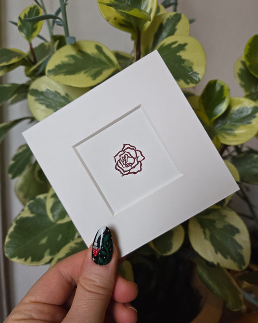 White card with a rose design held by a hand with decorative nails against a leafy background