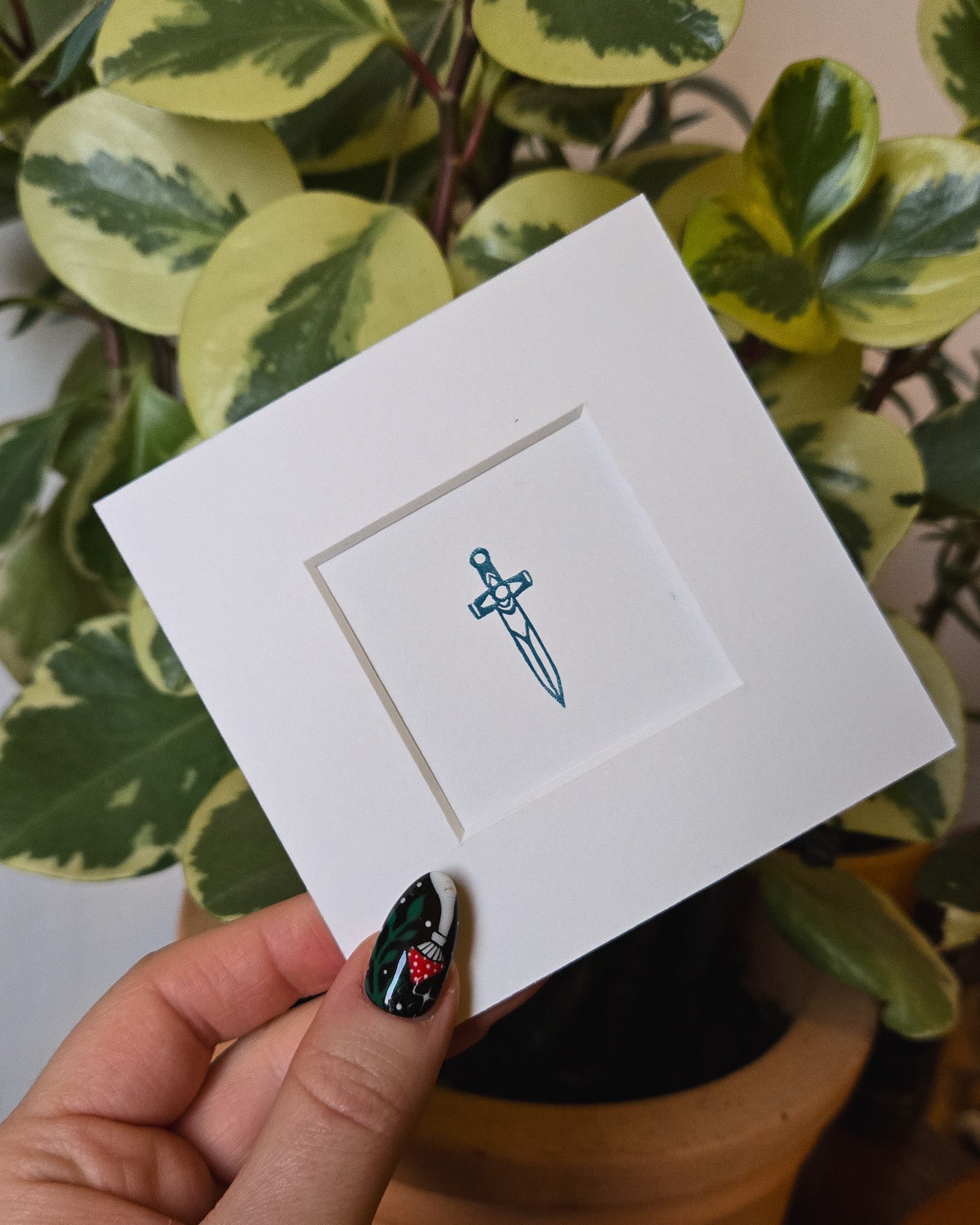 White card with a blue dagger linocut print held by a hand with green nail polish against a leafy background.