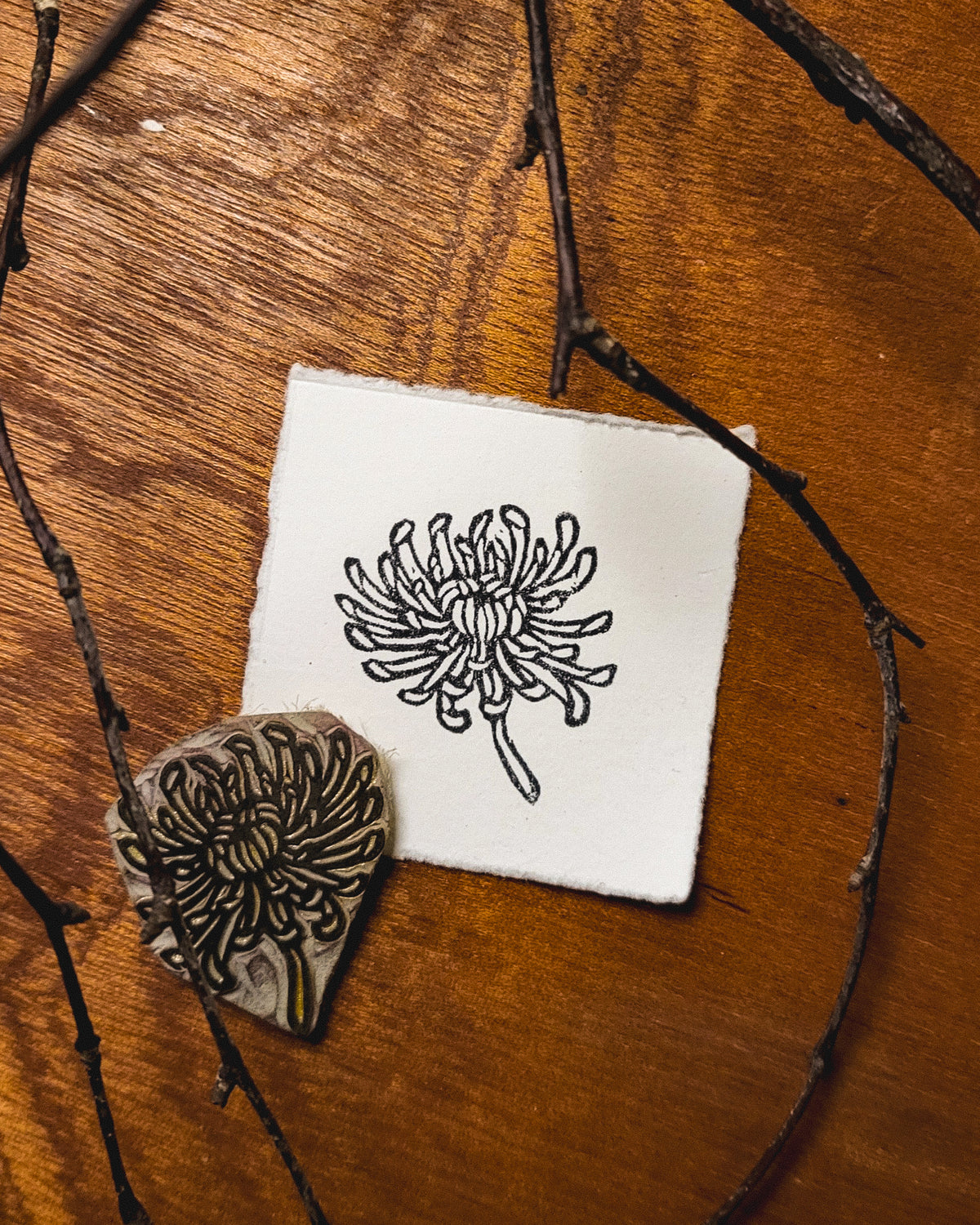 Wooden stamp with chrysanthemum floral design on a white paper square, surrounded by twigs on a wooden surface