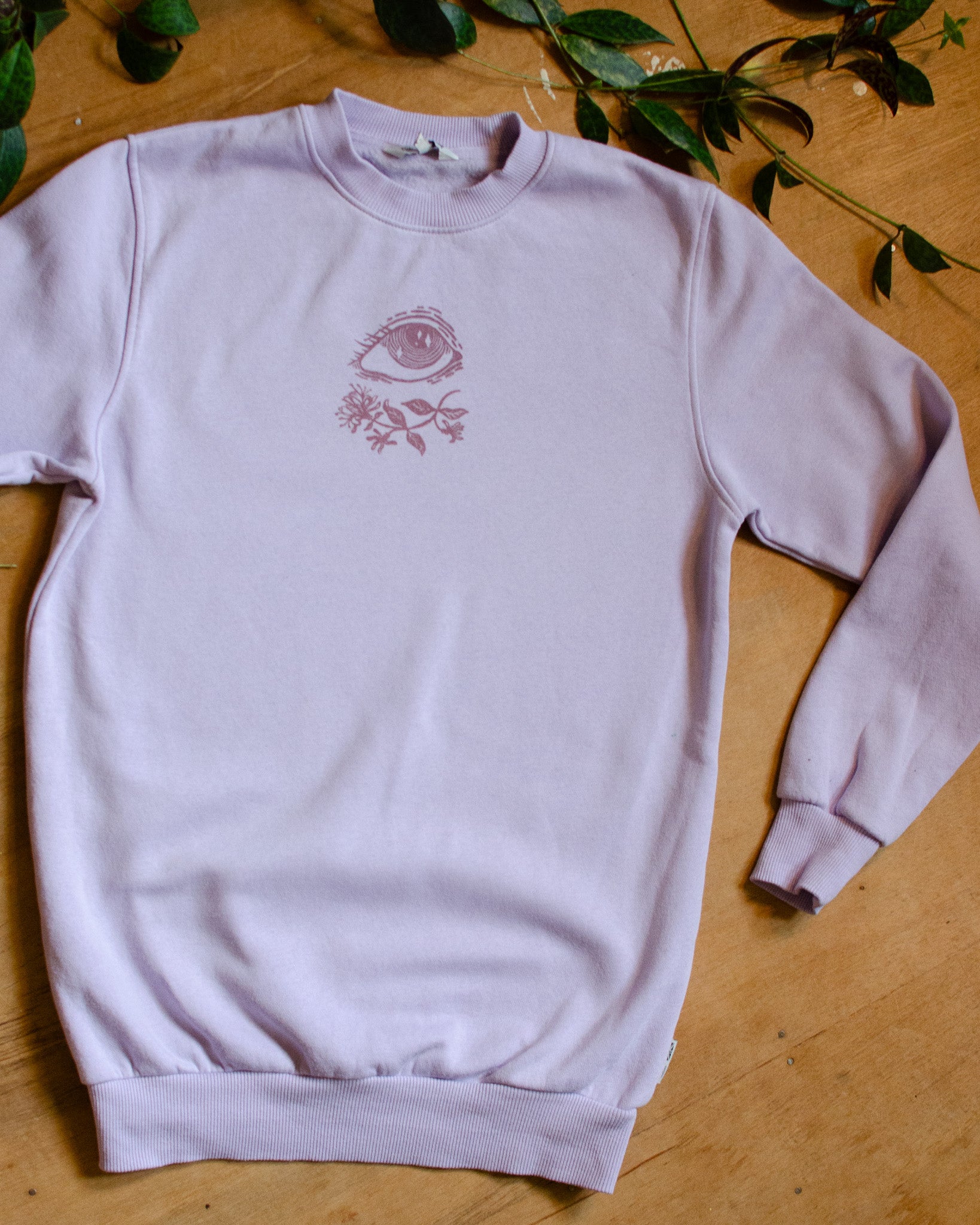 Light violet sweatshirt with a pink floral and eye design on a wooden surface with green leaves.