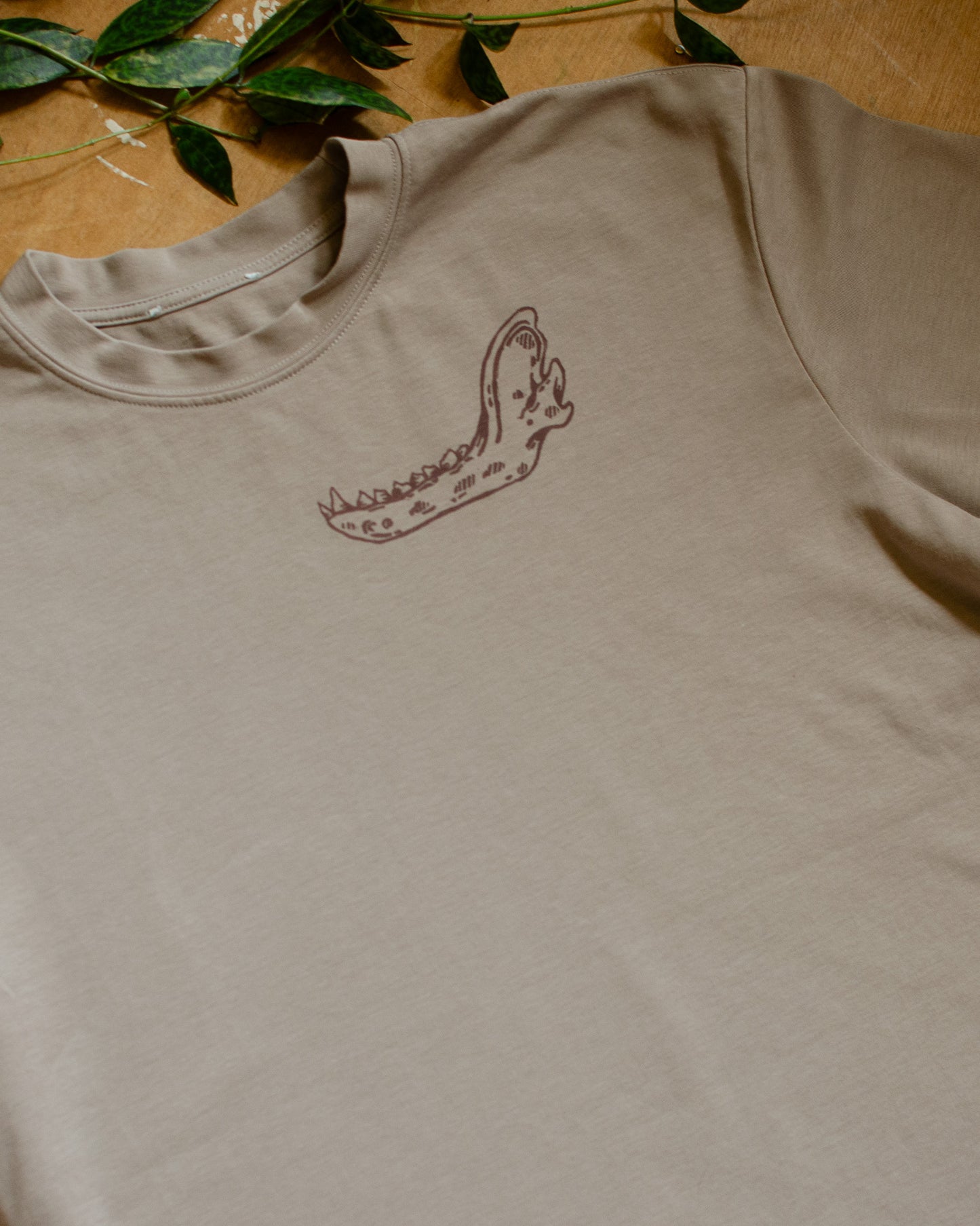 Beige t-shirt with an animal jawbone print on a wooden surface with green leaves.