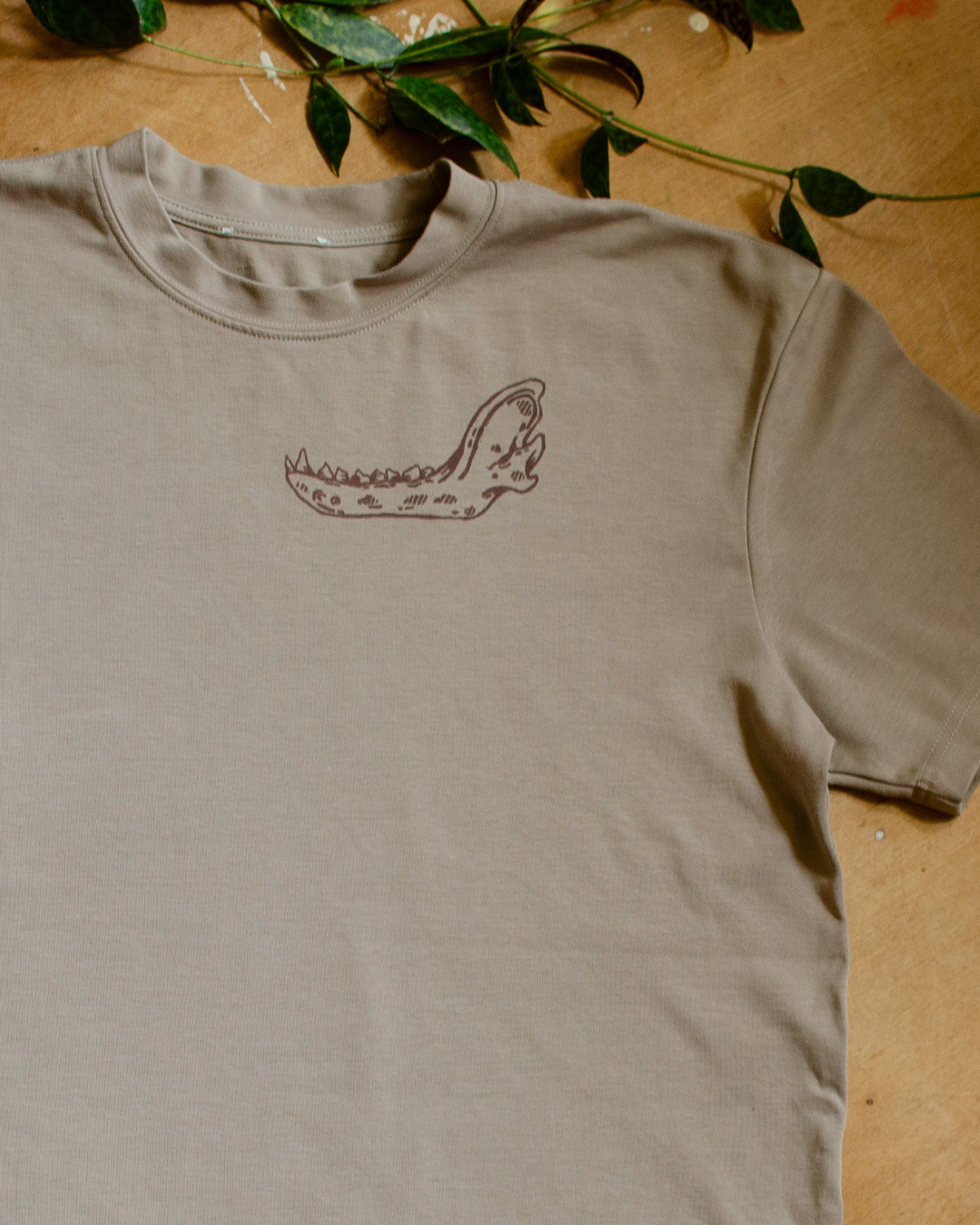 Beige t-shirt with an animal jawbone print on a wooden surface with green leaves.