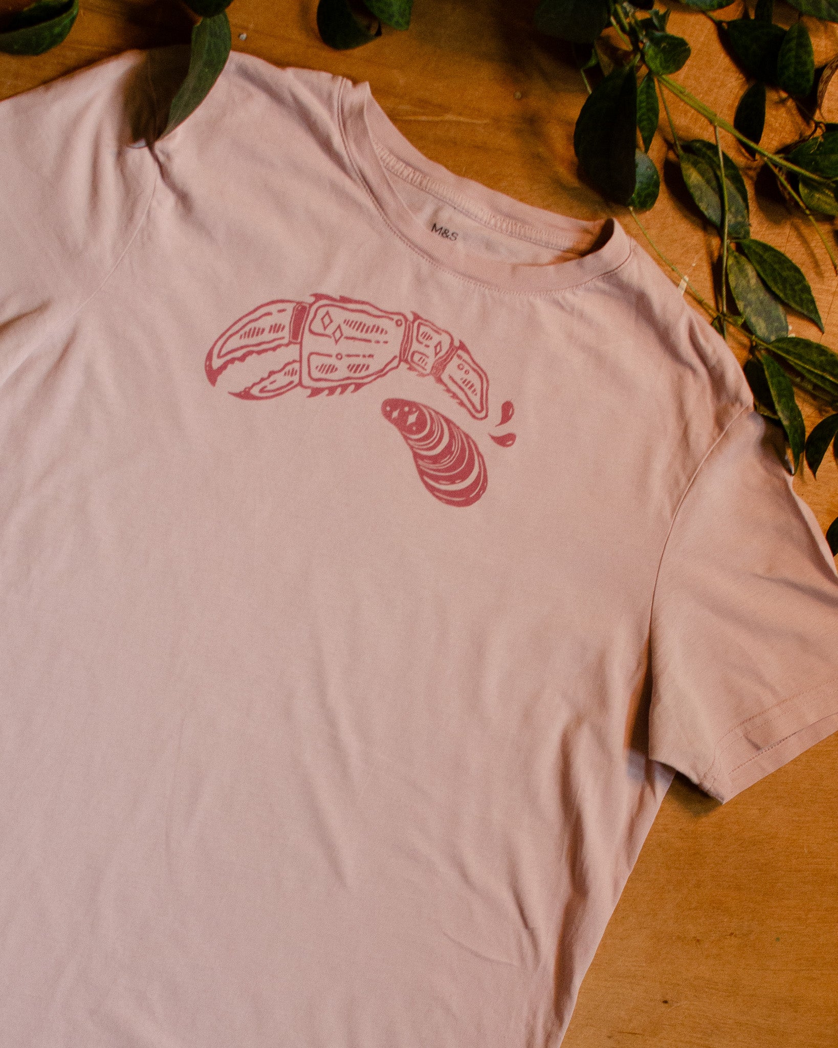 Pink t-shirt with dark pink lobster claw and mussel design on a wooden surface