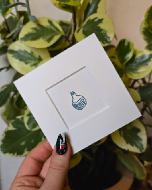 White card with a blue  potion bottle icon held by a hand with black nail polish against a leafy background
