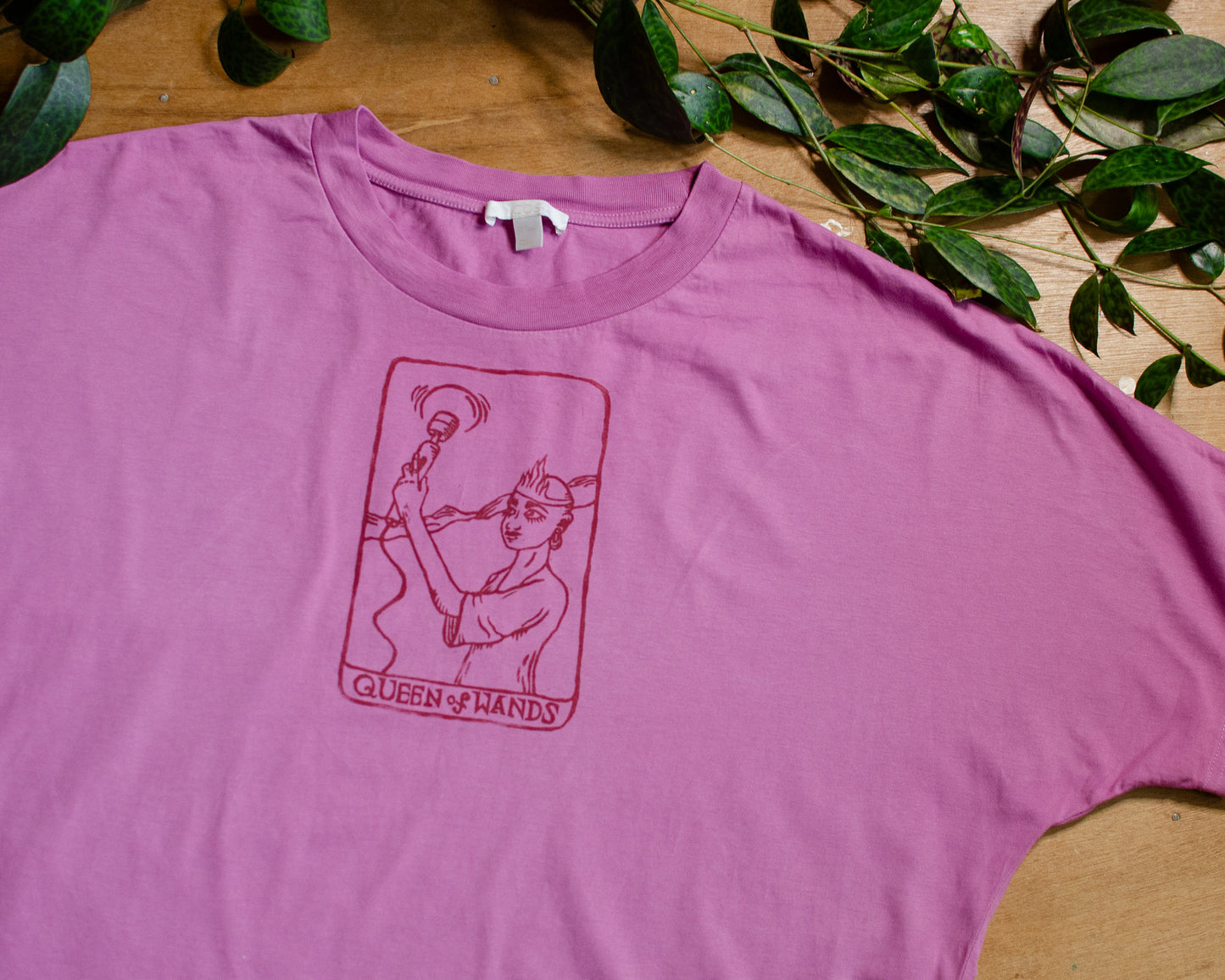 Pink t-shirt with a queen of wands tarot card design on a wooden surface with green leaves.