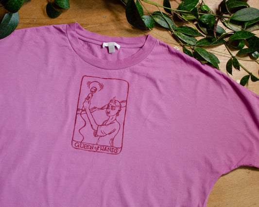 Pink t-shirt with a queen of wands tarot card design on a wooden surface with green leaves.