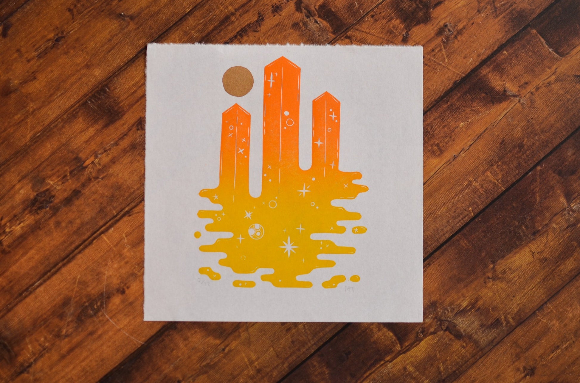Orange hand-printed abstract skyscrapers design on a white card with a gold circle, placed on a wooden surface