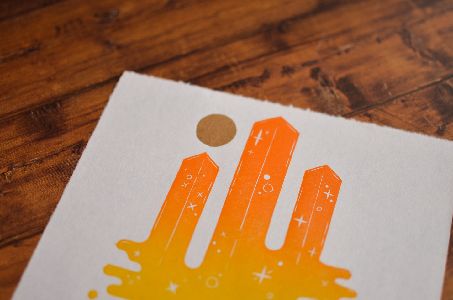 Orange hand-printed abstract skyscrapers design on a white card with a gold circle, placed on a wooden surface