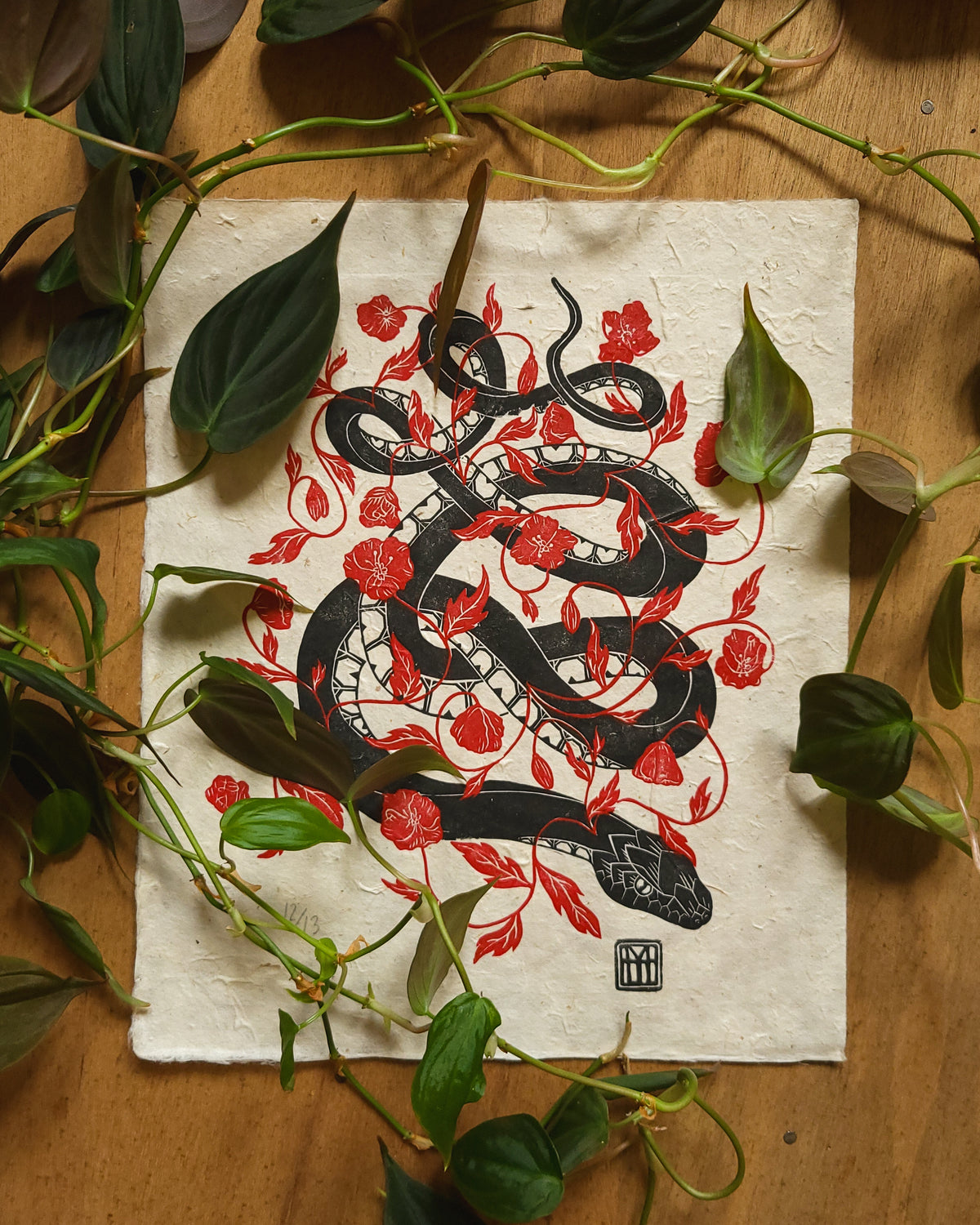 Linocut art print with a black snake and red flowers design on a wooden surface with leaves.