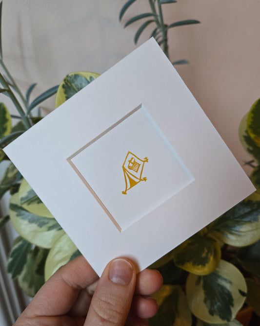 White card with a gold camping tent print held by a hand against a blurred green and white background