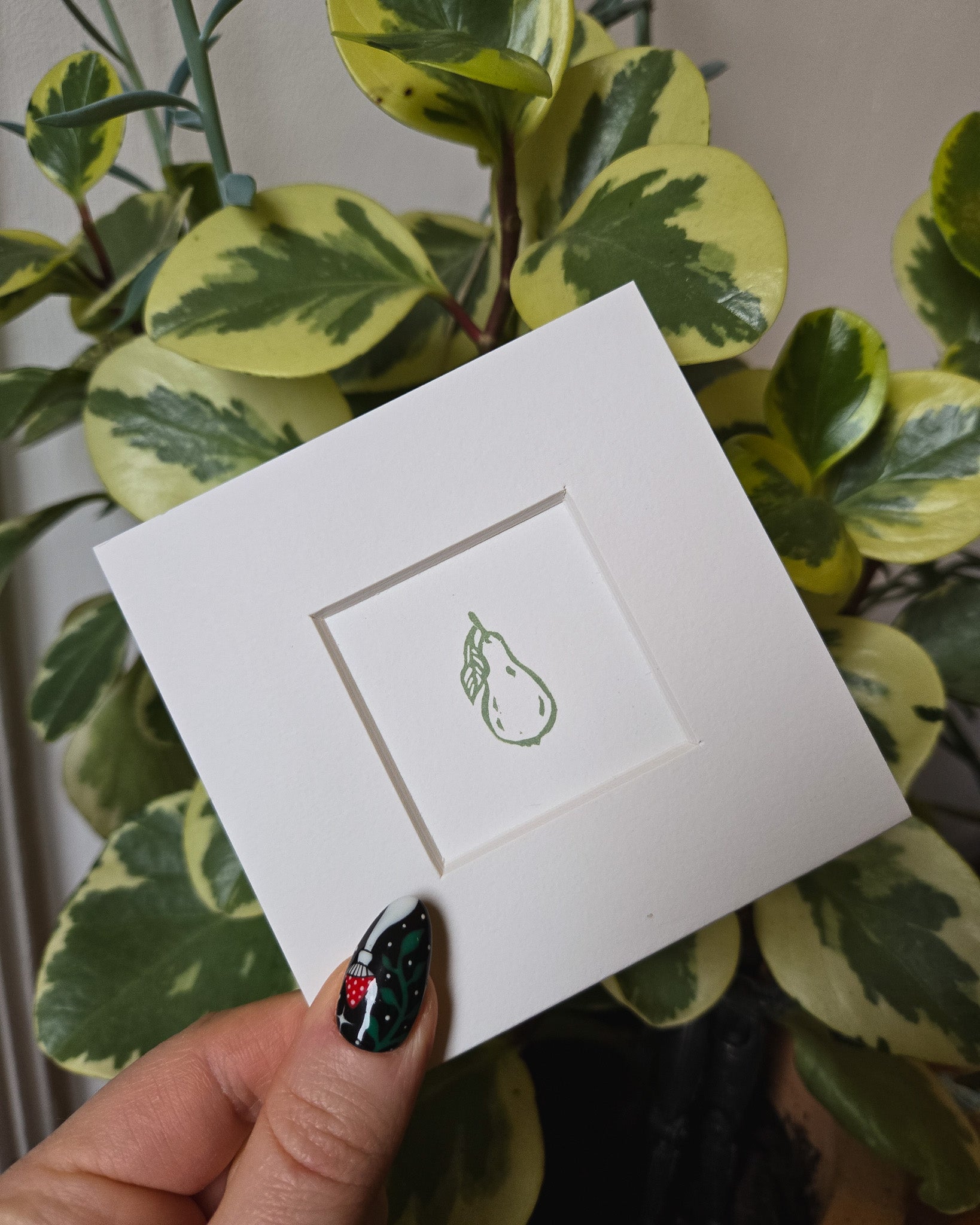 White card with a green pear design held by a hand in front of a plant.