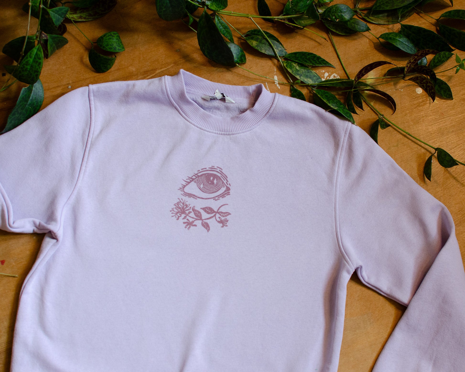 Light violet sweatshirt with a pink floral and eye design on a wooden surface with green leaves.
