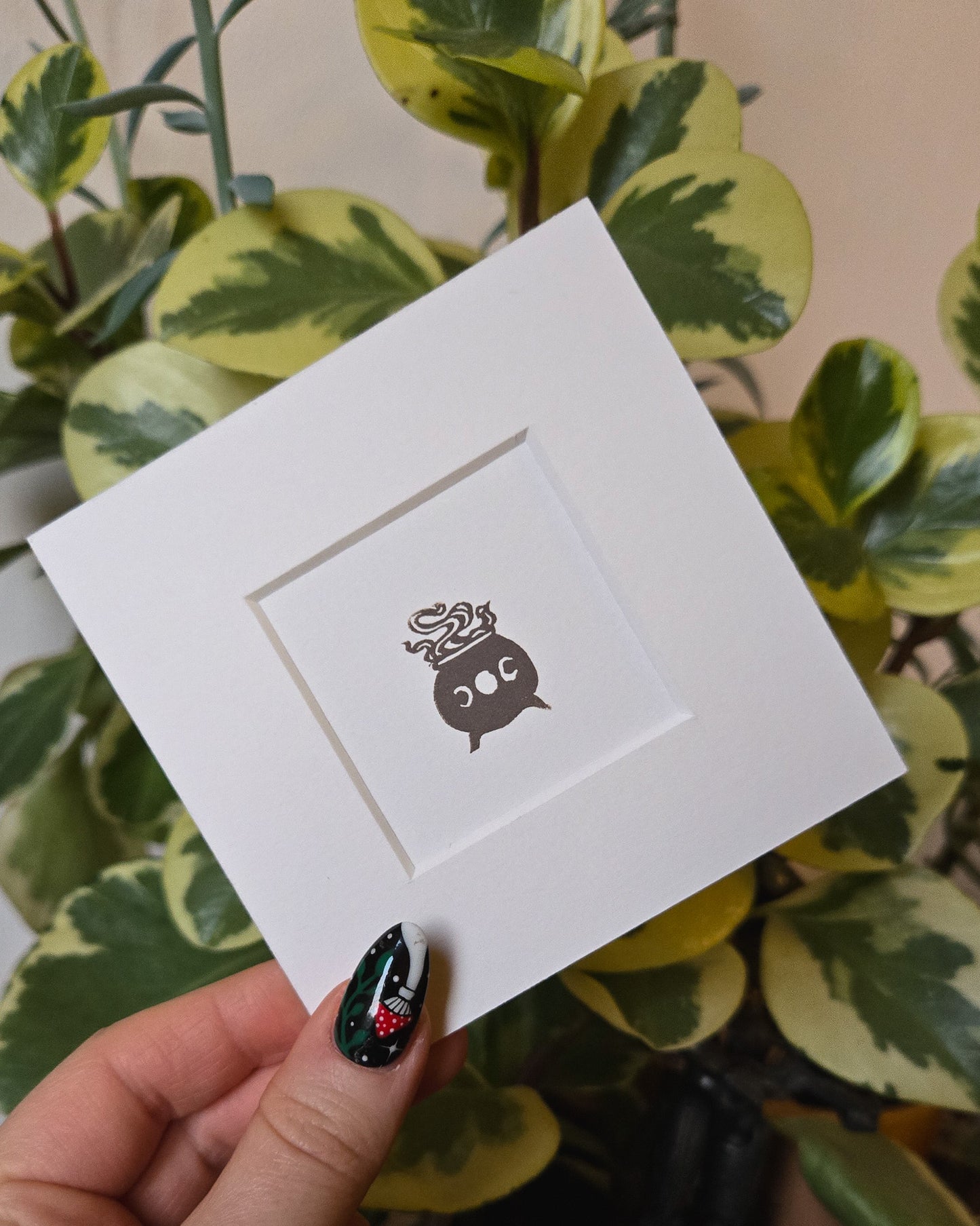 White card with a grey witchy cauldron design held by a hand in front of green plants