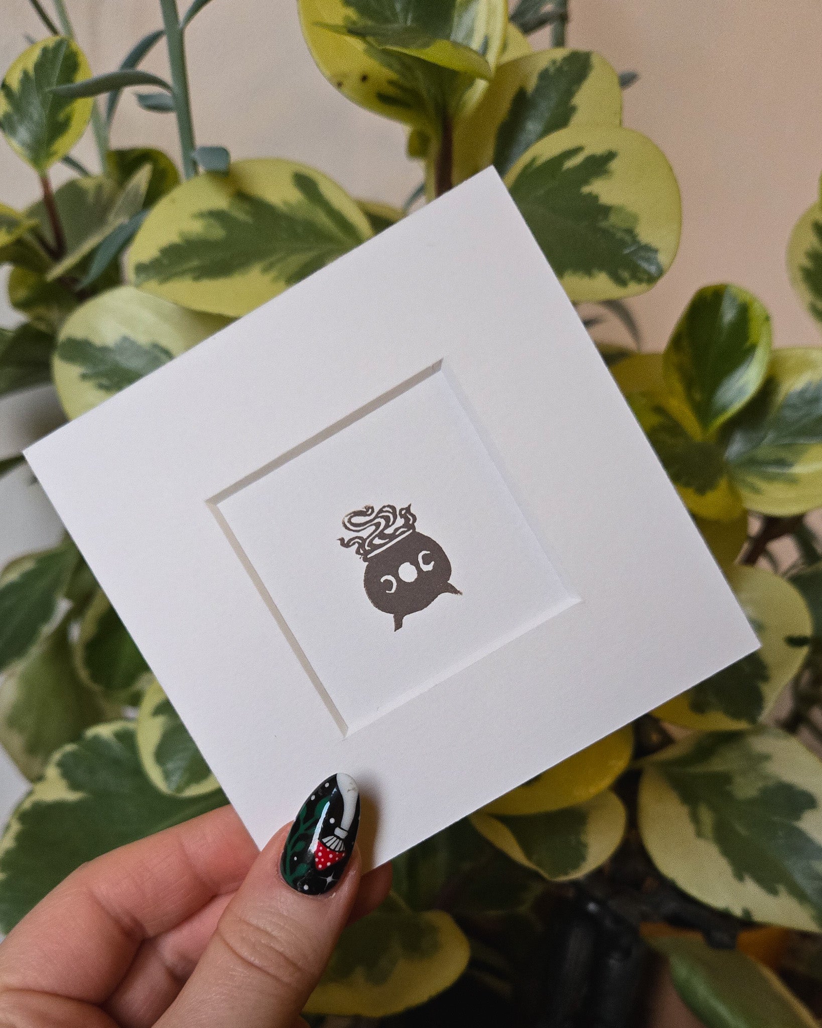 White card with a grey witchy cauldron design held by a hand in front of green plants