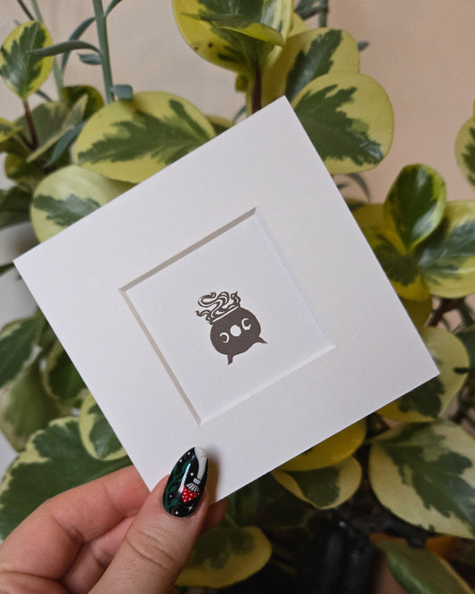 White card with a grey witchy cauldron design held by a hand in front of green plants