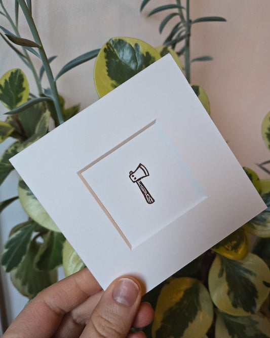 White card with a black axe symbol held by a hand in front of green foliage