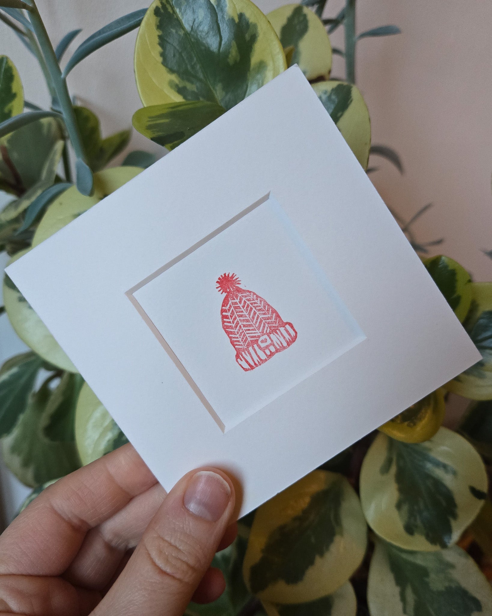 White card with a red hat design held by a hand against a leafy background